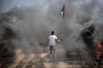 Un uomo cammina tra il fumo della guerra, tenendo in mano una bandiera palestinese.
