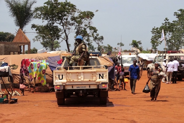 A Blue Helmet UN peacekeeper sits atop a UN vehicle patrolling the town of Bria, while civilians walk around.  