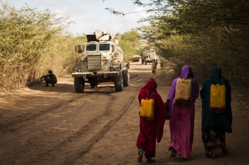 Donne trasportano l’acqua accanto a soldati alla ricerca di ordigni esplosivi